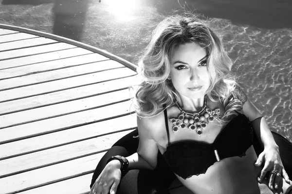 Black and white photo of beautiful sexy blonde woman laying near the swimming pool in black suite and bracelet and colorful beads