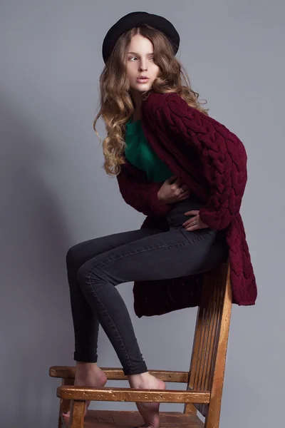 Fashion portrait of young beautiful woman model in casual wear (knitted cardigan, jeans and a green t-shir). without boots, in hat and natural make up on her face sitting on chair. Copy space.