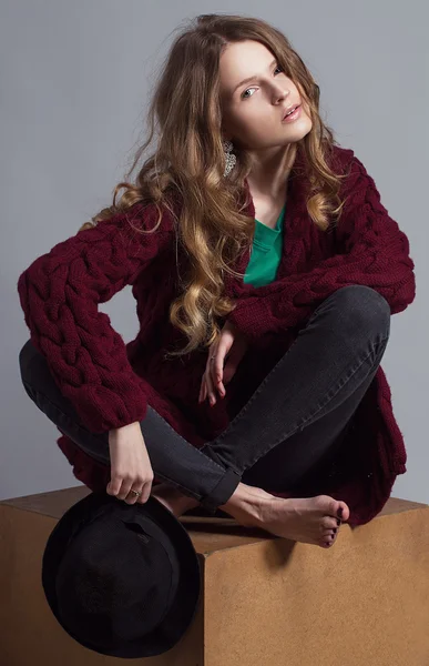 Fashion portrait of young beautiful woman model in casual wear (knitted cardigan, jeans and a green t-shir). without boots, in hat and natural make up on her face sitting on a wooden cube. Copy space.
