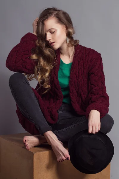 Fashion portrait of young beautiful woman model in casual wear (knitted cardigan, jeans and a green t-shir). without boots, in hat and natural make up on her face sitting on a wooden cube. Copy space.
