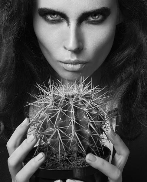 Sexy Beauty Girl with natural Lips and arty black make up on her eyes. Provocative Make up. Luxury Woman with Blue Eyes. Fashion Brunette Portrait on grey background holding a green cactus. Gorgeous Woman Face. Long Hair