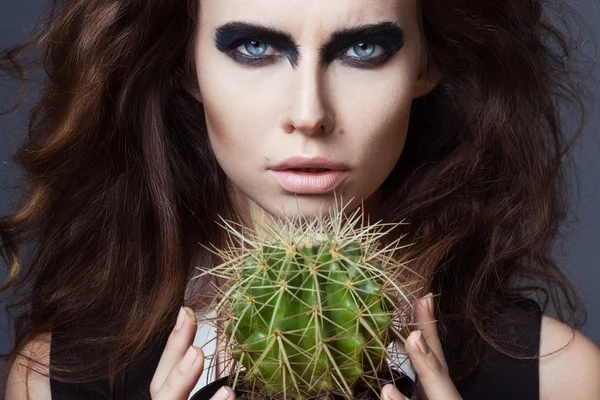 Sexy Beauty Girl with natural Lips and arty black make up on her eyes. Provocative Make up. Luxury Woman with Blue Eyes. Fashion Brunette Portrait on grey background holding a green cactus. Gorgeous Woman Face. Long Hair