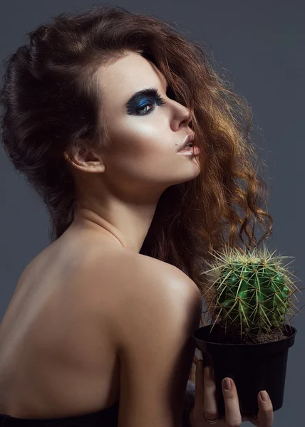 Sexy Beauty Girl with natural Lips and arty black make up on her eyes. Provocative Make up. Luxury Woman with Blue Eyes. Fashion Brunette Portrait on grey background holding a green cactus. Gorgeous Woman Face. Long Hair