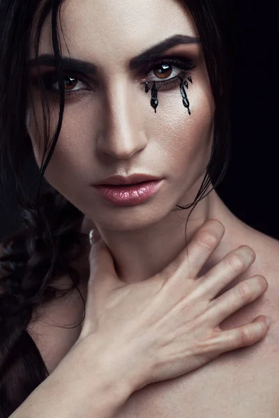 A photo of beautiful girl with perfect art makeup , painted black tears on her cheek and holding her hand on her  neck. Mystical and mysterious portrait