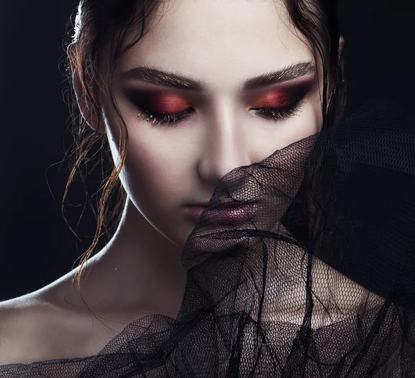 Portrait of mysterious beautiful brunette through black veil and posing over black background. Gold eyebrows, wet lips, red shadows. Close up. Vogue style. Studio shot. Haute couture concept.