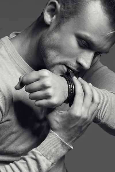 Advertising wrist watch concept. Beautiful (handsome) muscular male model with perfect body in grey jumper. He bites and unfastens the bracelet from the clock. Street style. Black and white, monochrome studio shot