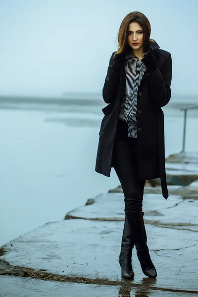 High fashion concept. Emotive portrait of beautiful brunette with long curly hair and perfect make up wearing black coat. Windy and misty weather.