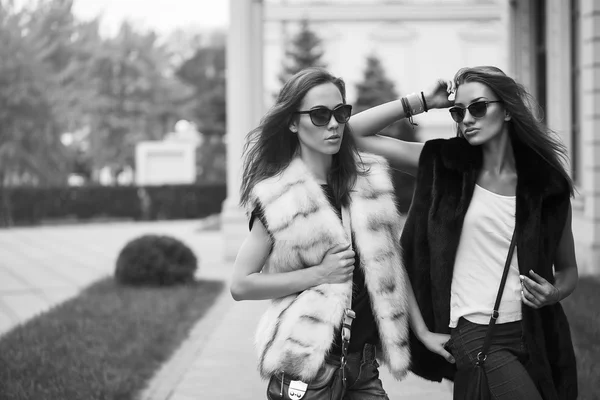 Fashion shot of two elegant beautiful girls in the sunset wearing sunglasses, fur vests . Two young women outdoor on the street. Shopping inspiration. Black and white concept