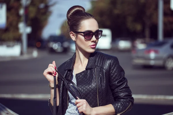 Female beauty concept. Portrait of fashionable young girl in casual black jeans, black jacket, white crop-top, sunglasses and small  bag posing on the street. Perfect hair & skin. Vogue style. outdoor shot