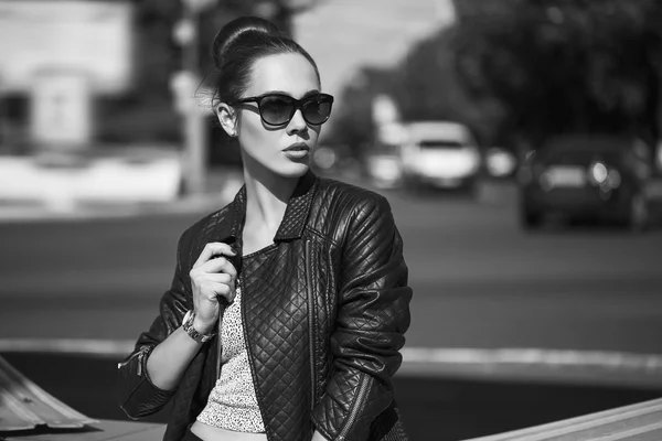Female beauty concept. Portrait of fashionable young girl in casual black jeans, black jacket, white crop-top, sunglasses and small  bag posing on the street. Perfect hair & skin. Vogue style. outdoor shot