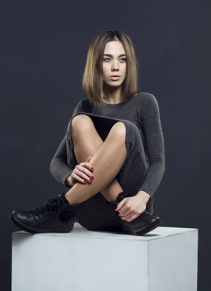 Sexy girl with brunette hair in grey dress sitting on white cube. Perfect hairdo and skin.