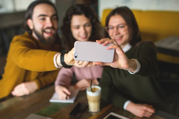 Three happy young friends taking selfie with smartphone in cafe
