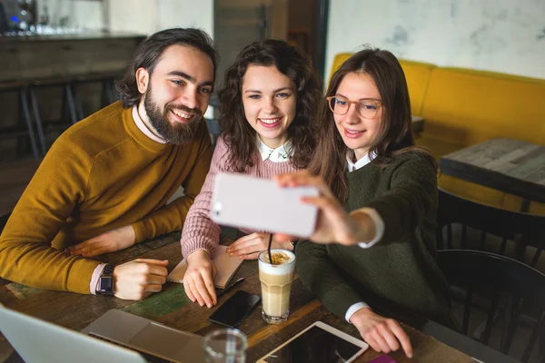 Three happy young friends taking selfie with smartphone in cafe