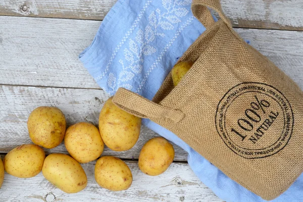 Organic fresh uncooked potatoes in a jute bag on a old weathered wooden white shelves background with vintage blue kitchen tea towel with empty copy space