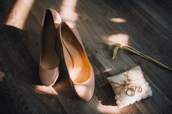 Beige bridal shoes laying on wooden floor with ears of wheat and pillow with wedding rings on side