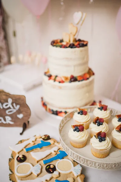 White wedding cake with cream decorated with strawberries and blueberries costs on a table and two wooden hearts on sticks with cupcakes