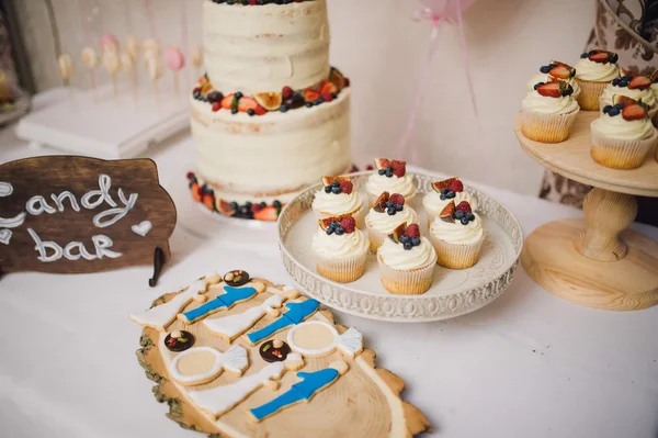 White wedding cake with cream decorated with strawberries and blueberries costs on a table and two wooden hearts on sticks with cupcakes