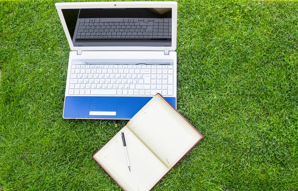 White Laptop, notebook and pen Placed On Green Grass Field.