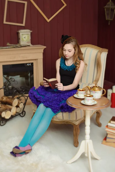 Young girl in the image of Alice in Wonderland is sitting in a chair near the fireplace and reading a book