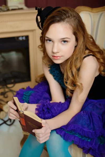 Young girl in the image of Alice in Wonderland is sitting in a chair near the fireplace and holds a book