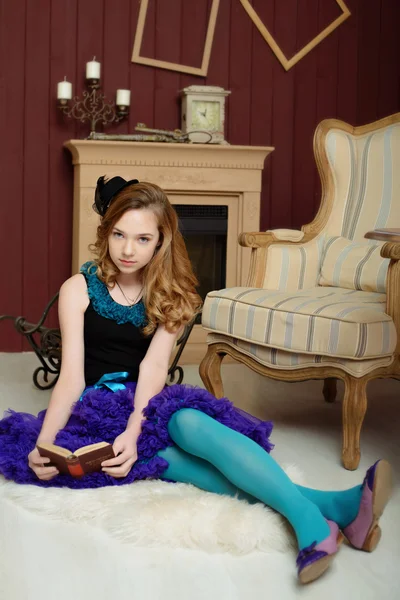 Young girl in the image of Alice in Wonderland is sitting near the fireplace and holds a book