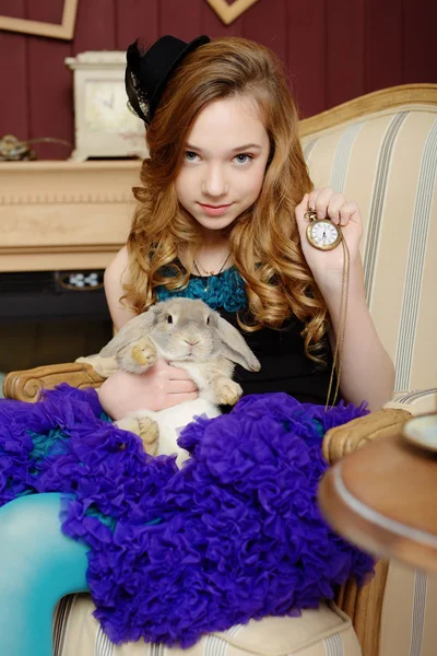 Young girl in the image of Alice in Wonderland is sitting in a chair by the fire, holds a rabbit and a pocket watches