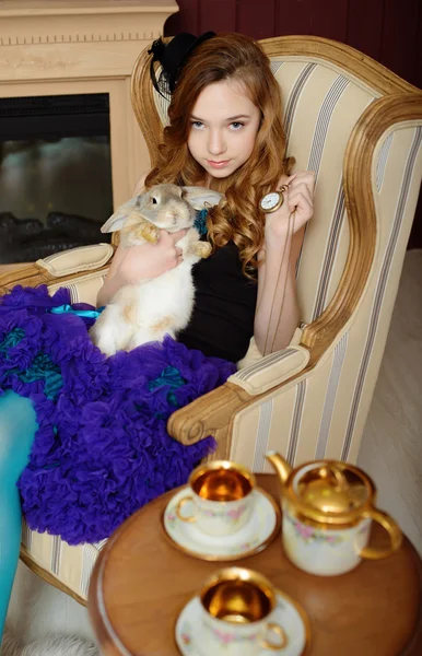 Young girl in the image of Alice in Wonderland is sitting in a chair by the fire, holds a rabbit and a pocket watches