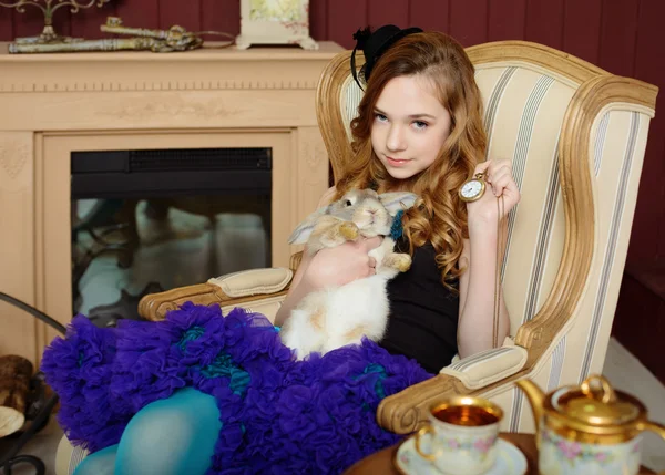 Young girl in the image of Alice in Wonderland is sitting in a chair by the fire, holds a rabbit and a pocket watches