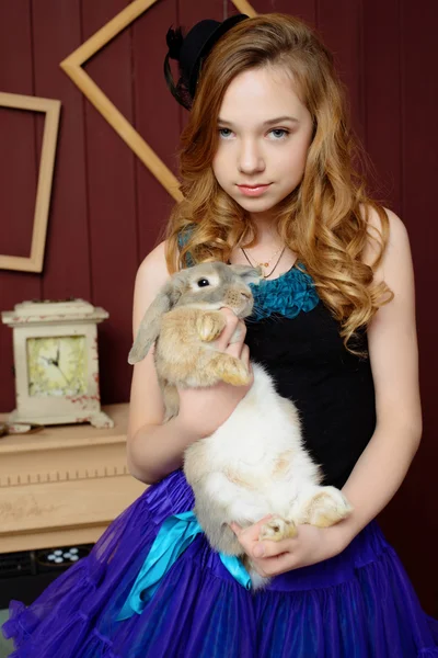 A young girl in the image of Alice in Wonderland stands near the fireplace and holds a rabbit