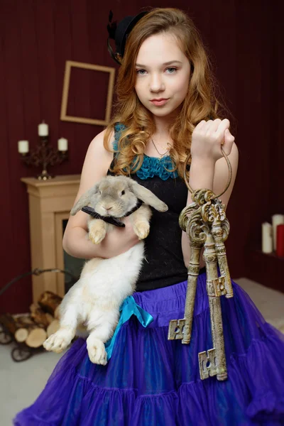 Young girl in the image of Alice in Wonderland stands near the fireplace and holds a rabbit and a bunch of keys