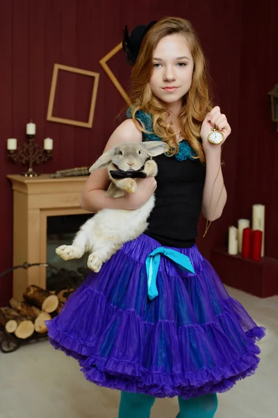 A young girl in the image of Alice in Wonderland stands near the fireplace and holds a rabbit and pocket watches