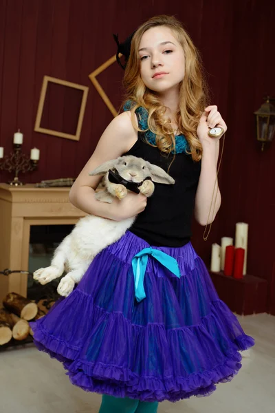 A young girl in the image of Alice in Wonderland stands near the fireplace and holds a rabbit and pocket watches