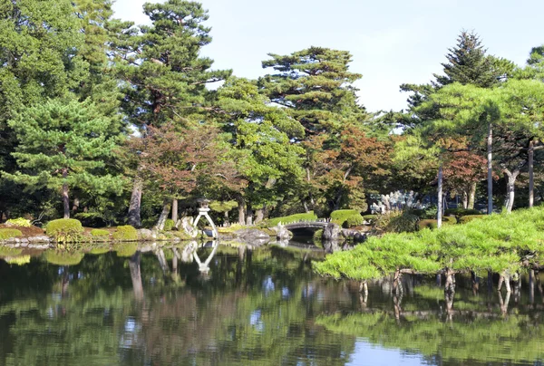 Japanese style garden park with stone bridge, lantern on a lake edge with green pine trees, autumn leaf trees and water reflections