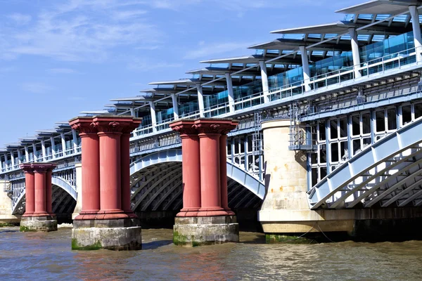 View from south bank of west side of arch wrough iron London Blackfriars Railway Bridge with platforms crossing River Thames in London along old red pillars, solar panels powered