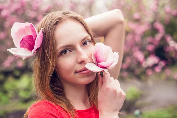 Cute girl with freckles smiling on a background of flowers. Girl holds in the hands of magnolia and looking at the camera. Close-up shot of spring.