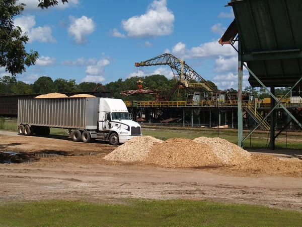 Pile of wood chips beside, filled semi truck alongside a lumberyard with logging crane working in the background.