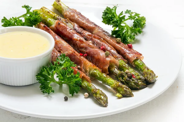 Grilled green asparagus wrapped in prosciutto bacon and hollandaise sauce, white plate on a white wooden table