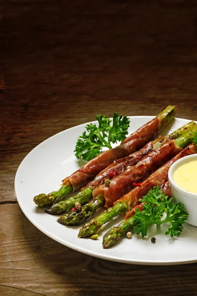 Grilled green asparagus wrapped in prosciutto bacon with parsley and hollandaise sauce on a dark rustic wooden background, view from above, copy space, vertical