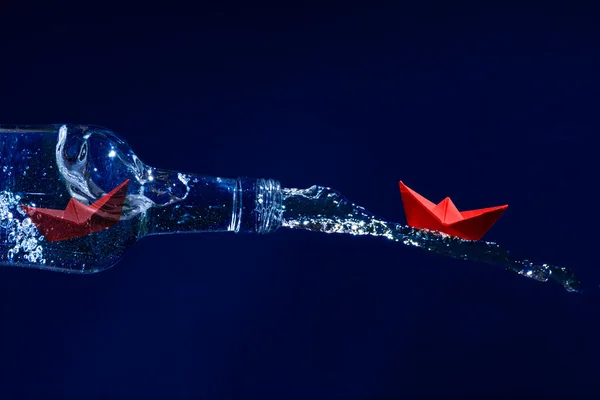 Red paper boats escape on a water splash from a bottle, dark blu