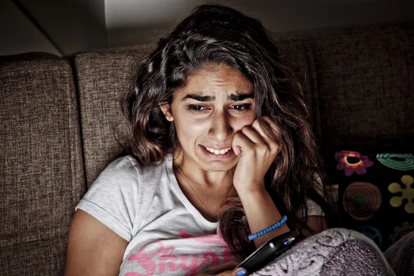 Scared young woman watching tv. Brunette girl sitting on couch with remote control