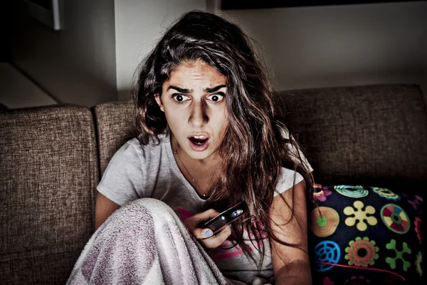 Scared young woman watching tv. Brunette girl sitting on couch with remote control