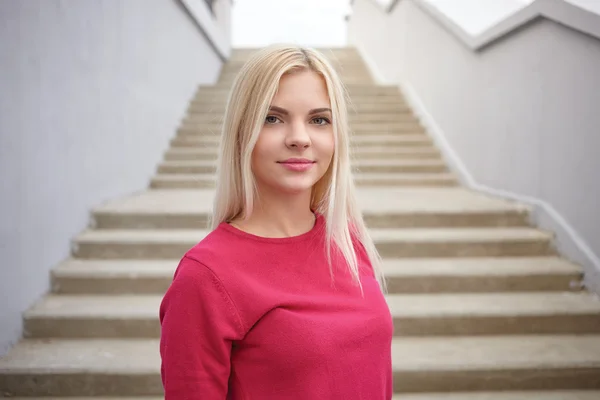 Close up portrait of young beautiful blonde woman with trendy makeup in fuchsia blouse against concrete stairs background