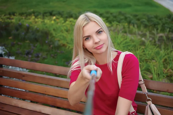 Young beautiful blonde woman with trendy bright makeup in fuchsia blouse pose faces doing a closeup selfie portraits sitting on the park bench