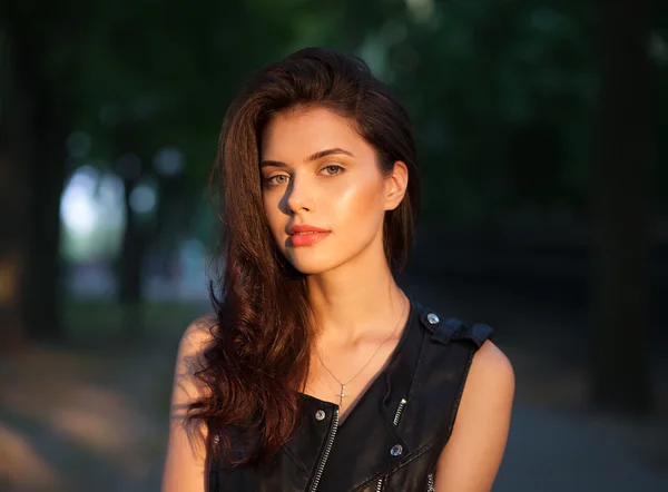 Closeup portrait of happy young beautiful brunette woman in black leather jacket posing on sunset outdoors  with blurry foliage background