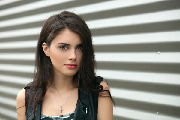 Closeup portrait of young beautiful brunette woman in black leather jacket posing outdoors against urban style background of metal strips