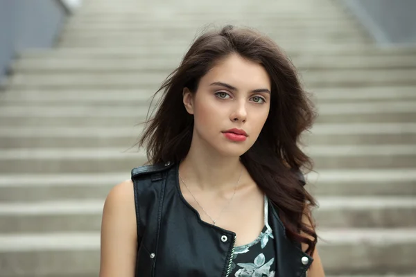 Closeup portrait of young beautiful brunette woman in black leather jacket posing outdoors against concrete stairway background