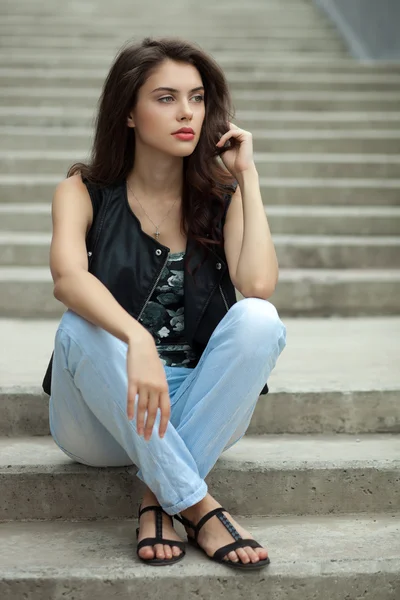 Young thoughtful fashionable brunette woman in black leather jacket posing sitting on concrete stairway
