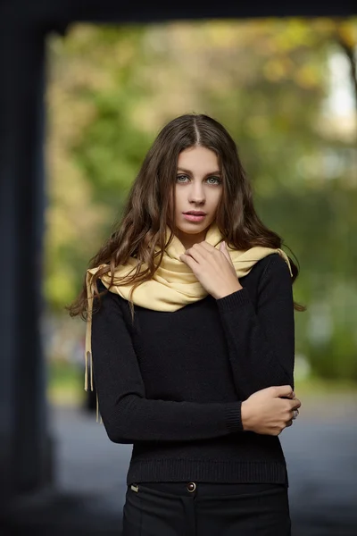 Young beautiful brunette woman with long wavy hair posing outdoors in black sweater and yellow scarf with blurry park background