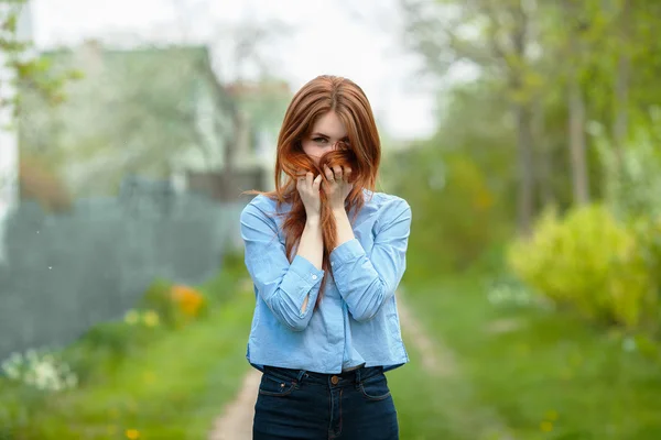 Beautiful healthy young redhead woman hides her face in her hair posing on the green grass of country road