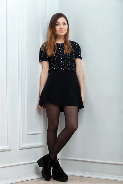 Young trendy dressed woman posing in black skirt boots tights shirt near white wall indoor